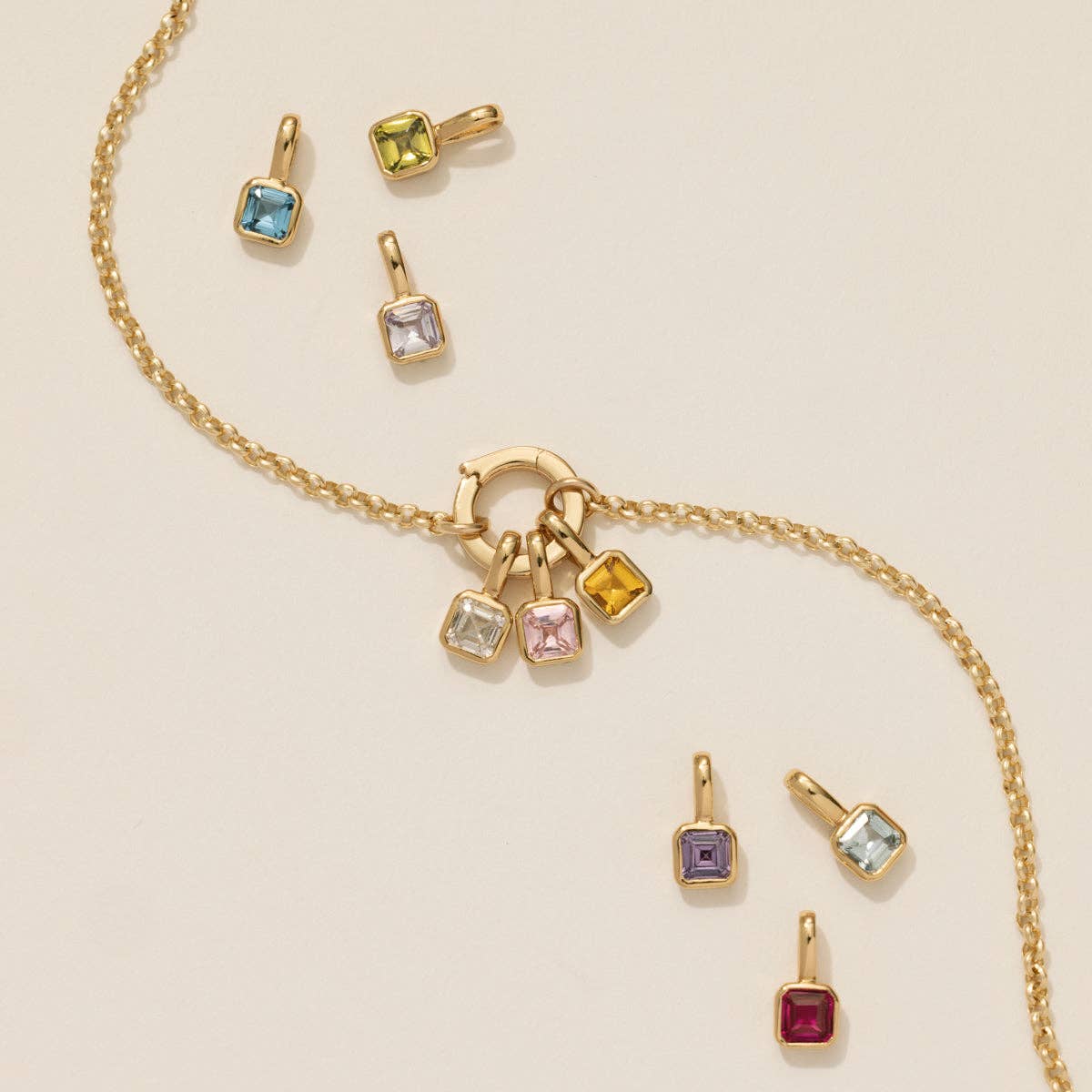 close up of the rolo circle lock charm necklace with three gemstone charms attached and six gemstone charms surrounding the necklace on a beige background