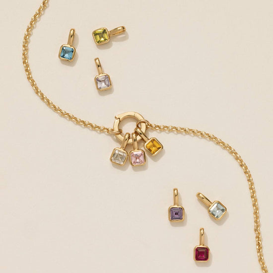 close up of the rolo circle lock charm necklace with three gemstone charms attached and six gemstone charms surrounding the necklace on a beige background