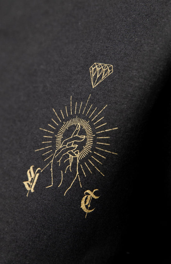 close up view of the front design on the tee, featuring a gold hand pointing up with gold like sun surrounding with a diamond on top and an old english a to the left and t to the right