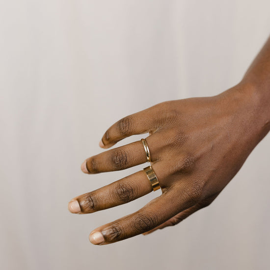 close up of a hand model wearing a large gold band on their right middle finger and a half round yellow gold band on their ring finger