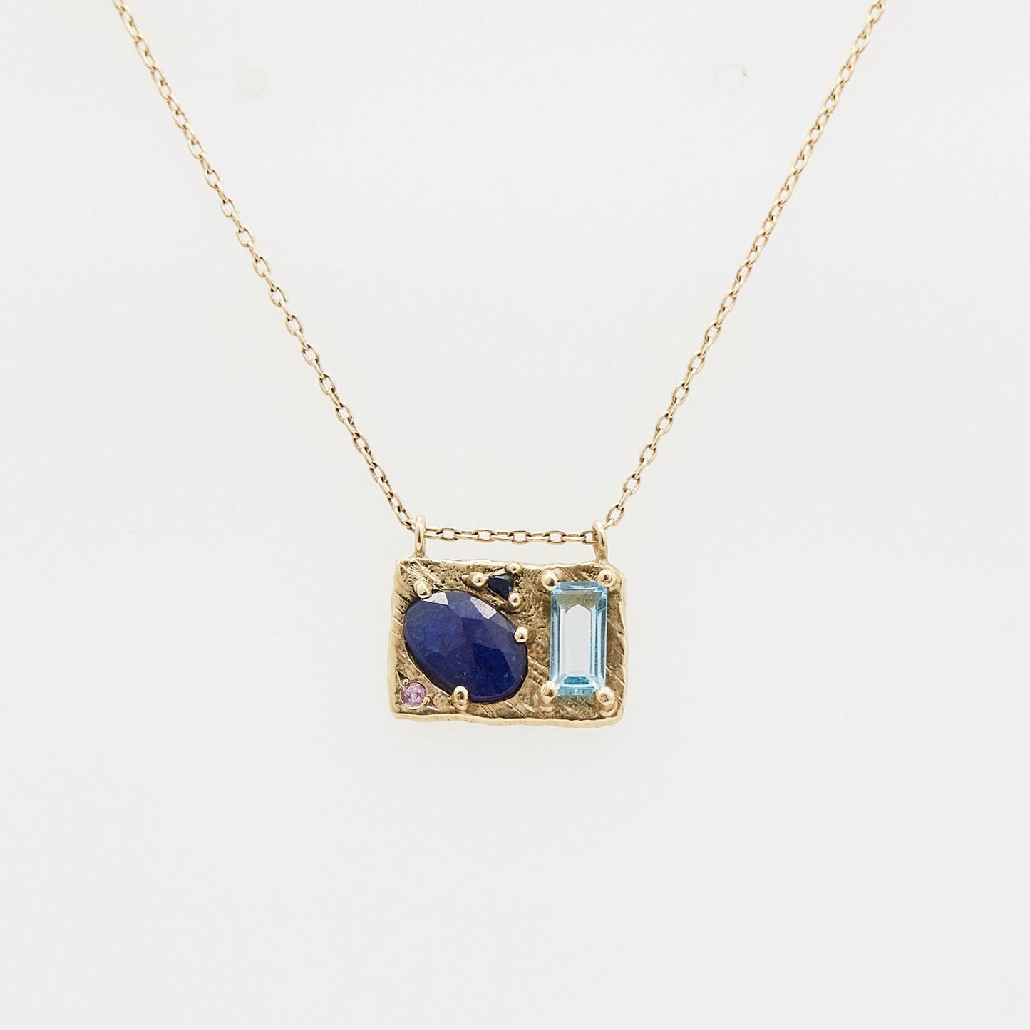 gold rectangle pendant necklace with oval blue stone, rectangle light blue stone, triangle blue stone and round pink stone on white background