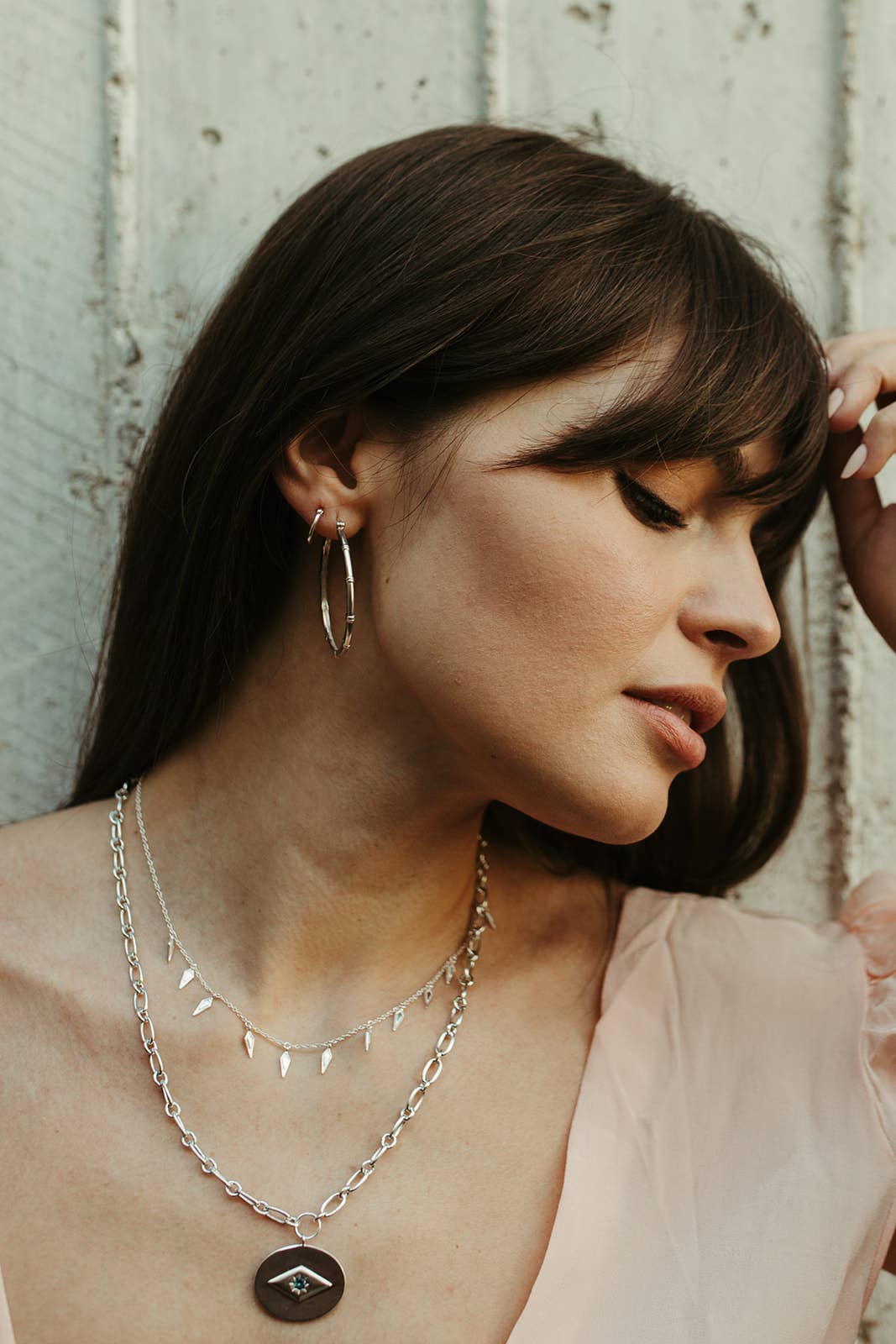 Woman wearing layered silver necklaces and hoop earrings against a wooden background