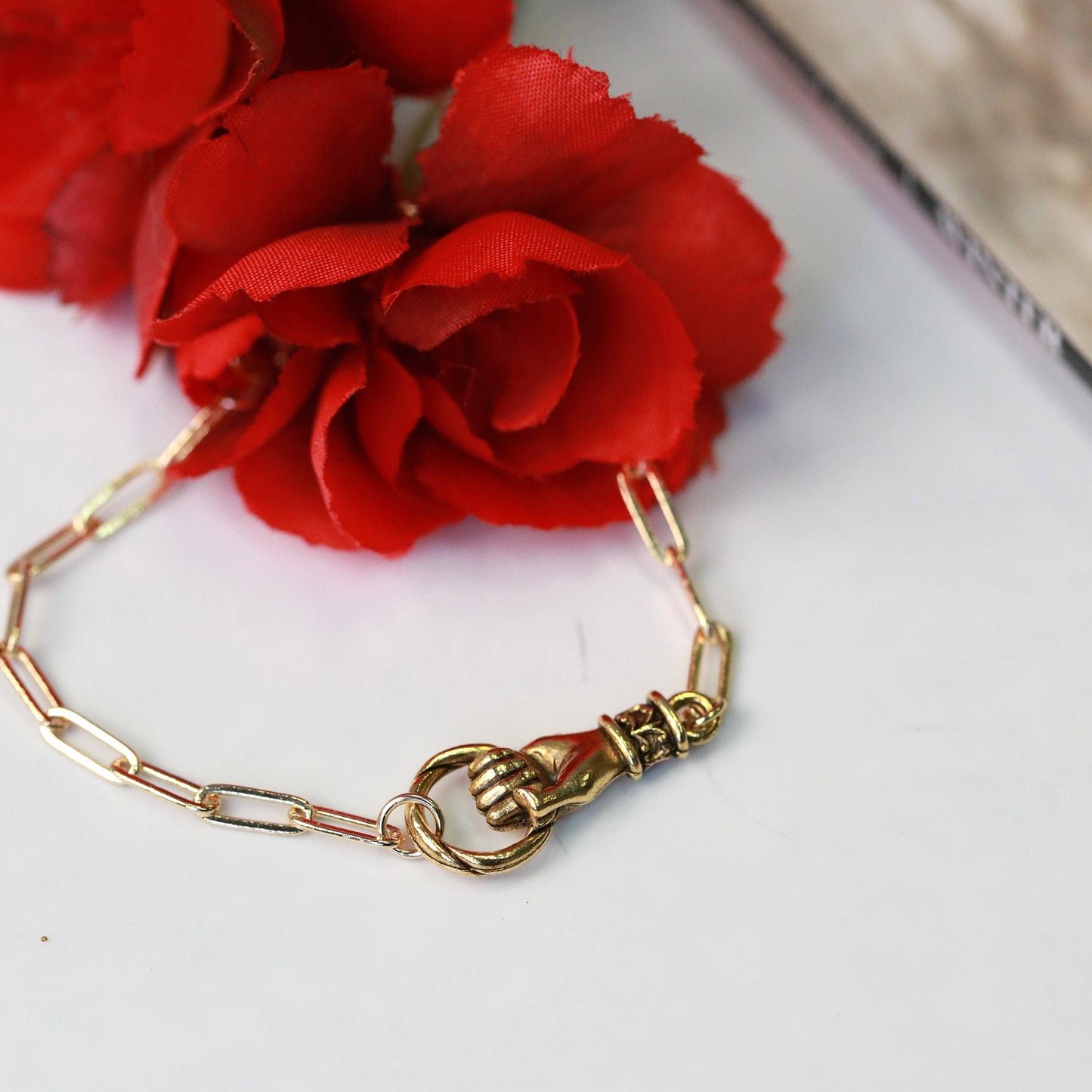 Gold bracelet with a hand holding a circle clasp on a white surface with red flowers.