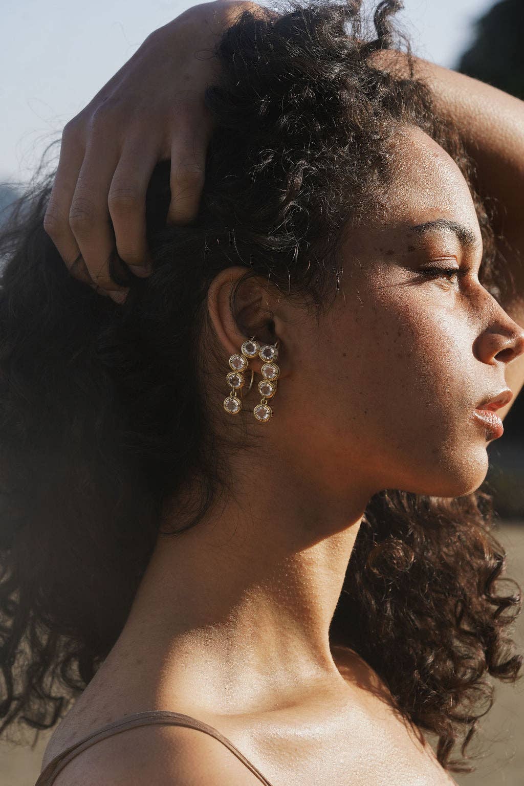 Woman with curly hair and earrings, with a hand on her head, against a blurred background