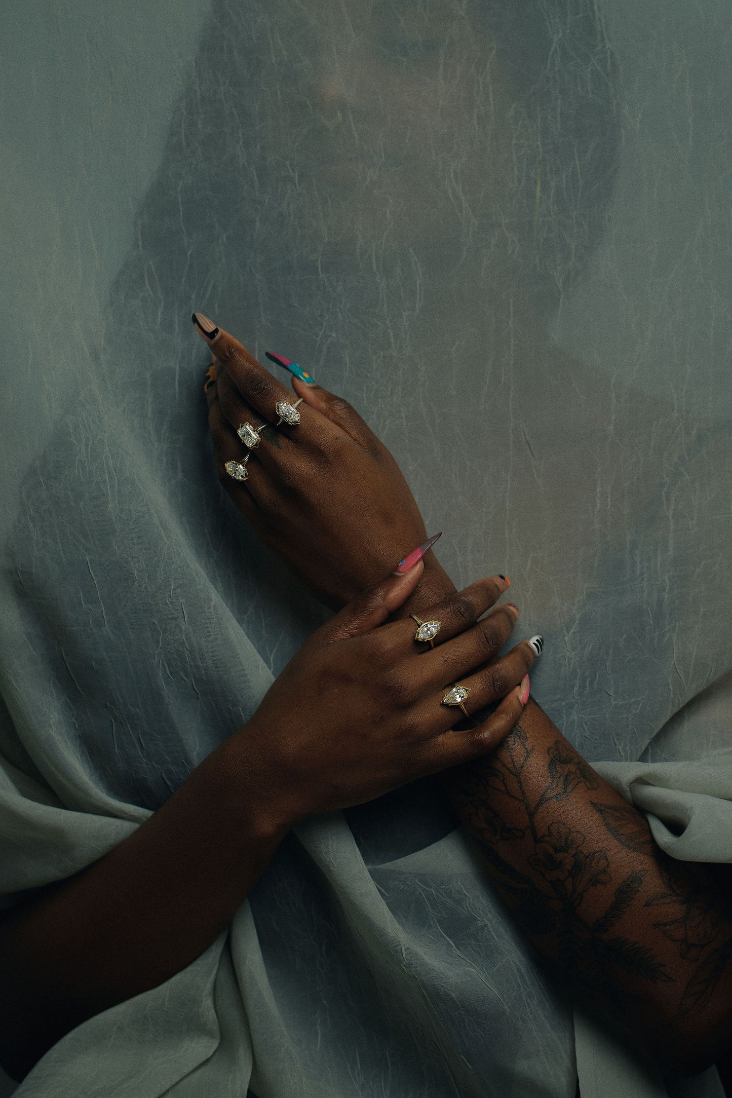 A close-up of a person's hands and arms adorned with multiple gold and diamond rings, resting on draped light blue-gray fabric.