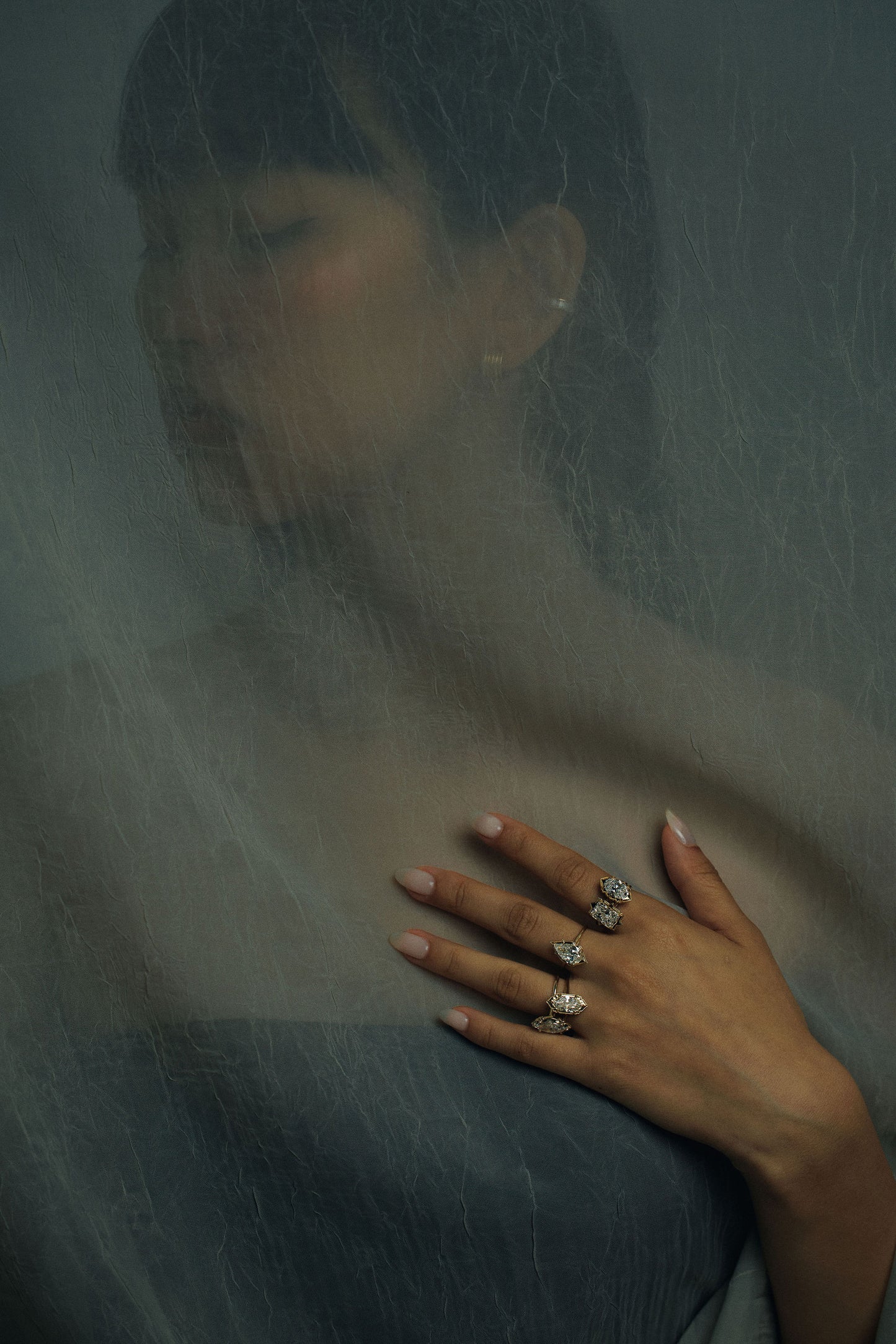 A close-up of a person's hand resting on their chest, showcasing several gold diamond rings. The background is a dark, moody gray with a sheer overlay, creating a soft, artistic look.