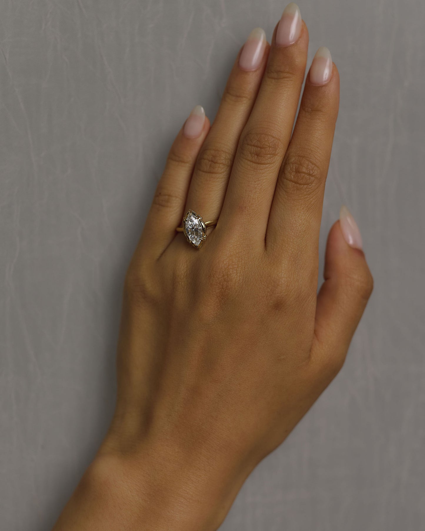 A close-up of a hand with manicured nails wearing a gold ring with a marquise-shaped white diamond, set against a textured gray background.