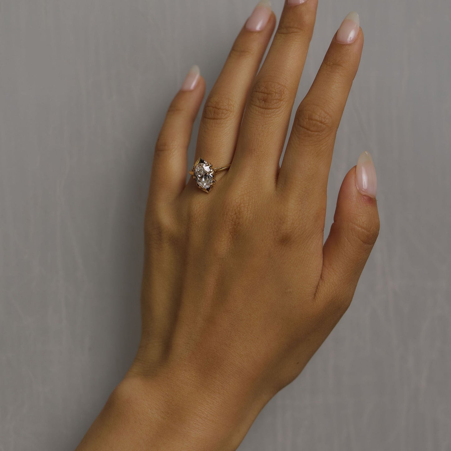 A close-up of a hand with manicured nails wearing a silver-toned ring with a clear central gemstone and small side stones, set against a plain gray background.