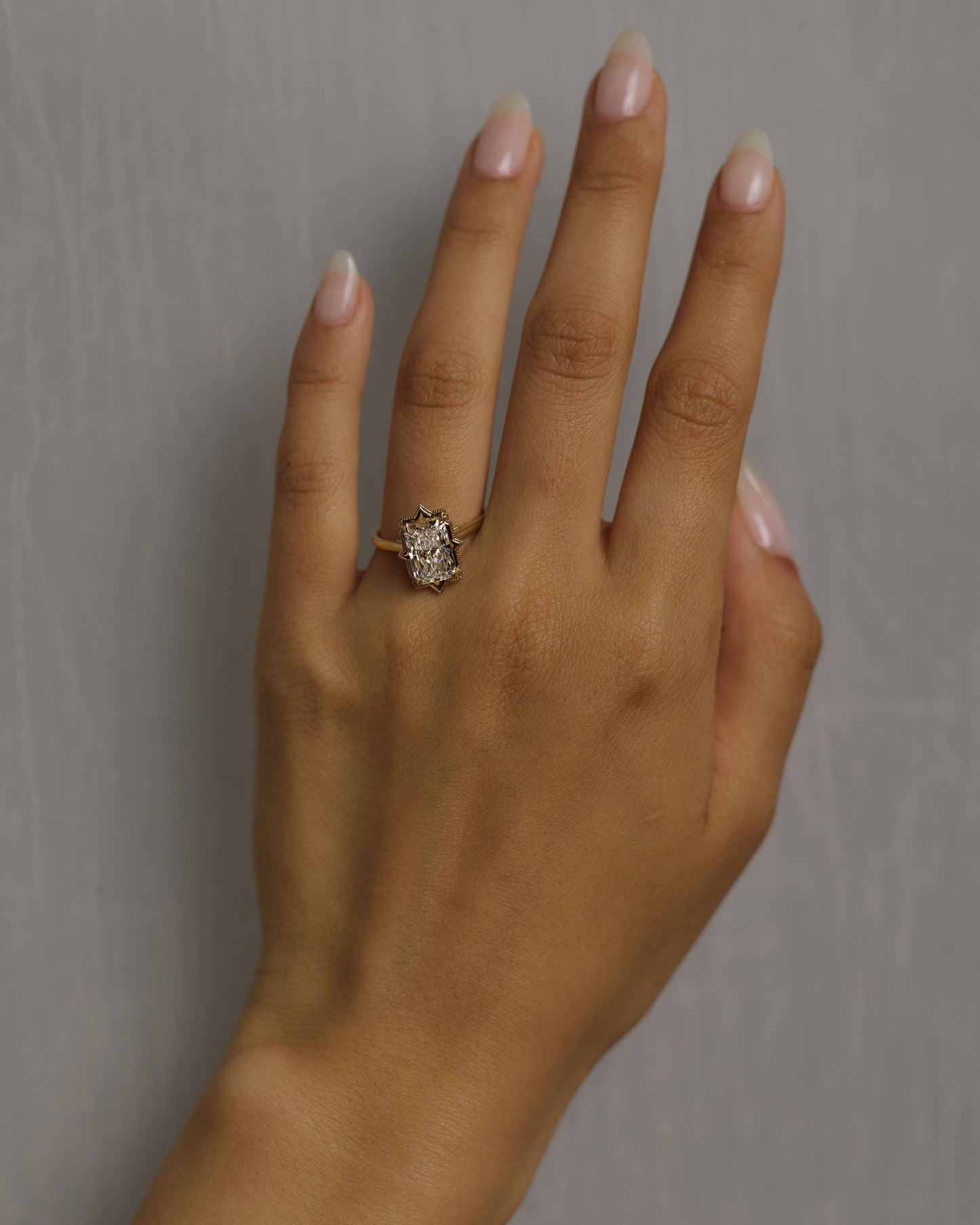 A close-up of a hand wearing a vintage-style gold ring with a large white diamond, set against a neutral gray background.