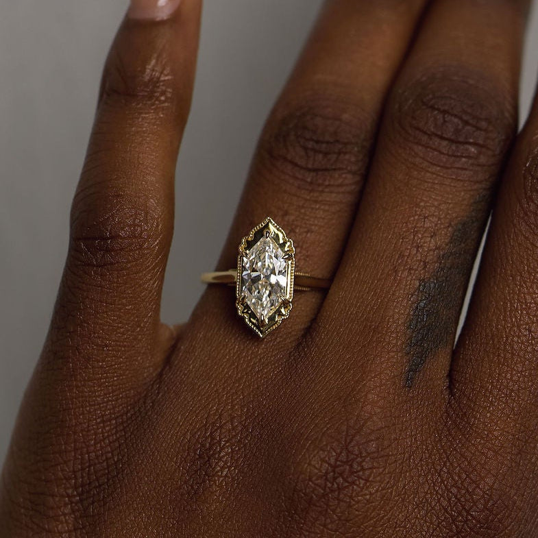 A close-up of a person's hand wearing a gold ring with a marquise-cut clear gemstone, set against a plain gray background.