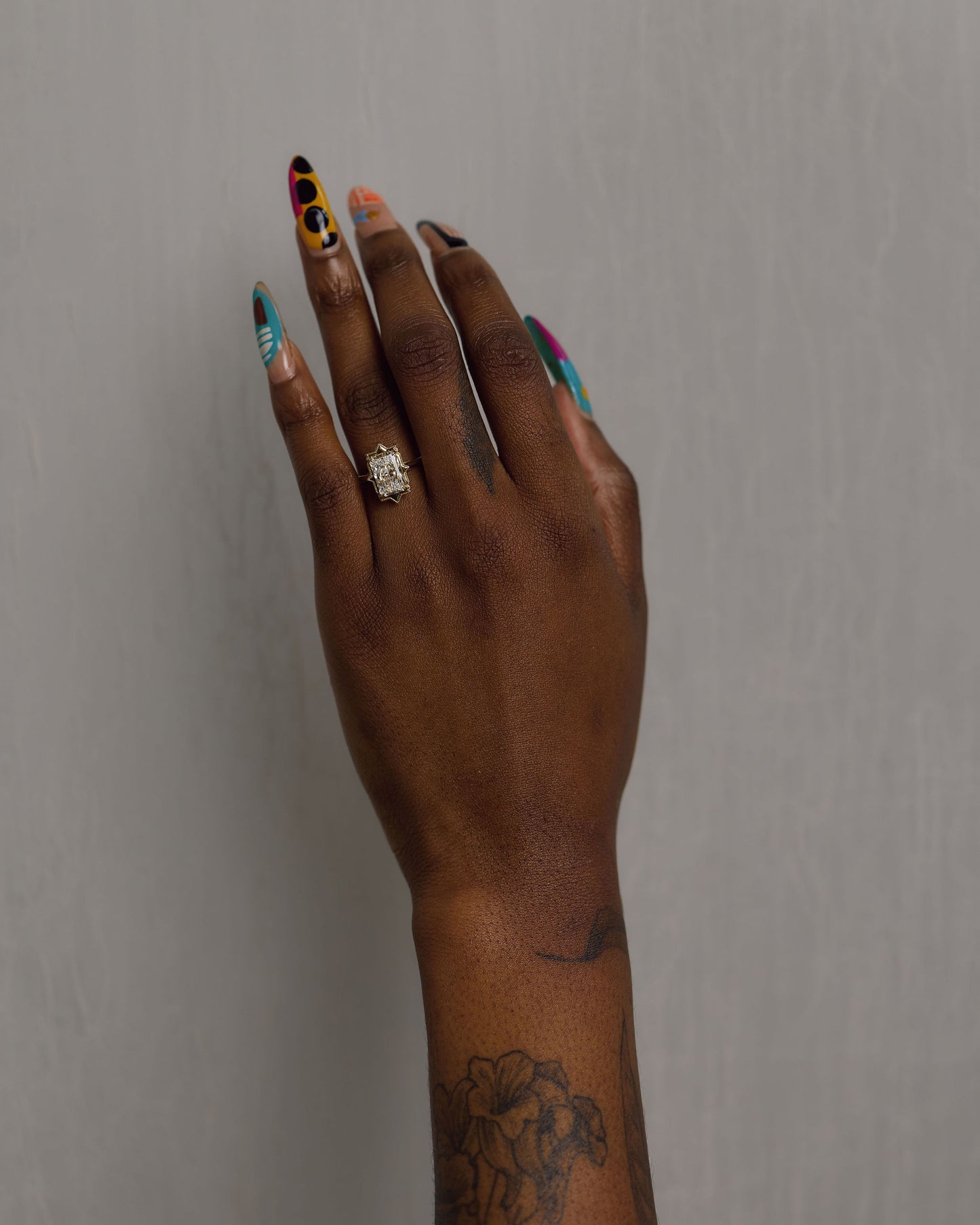 A close-up of a person's hand with long, colorful stiletto-style nails and a gold ring with radiant white diamond, posed against a plain gray background.