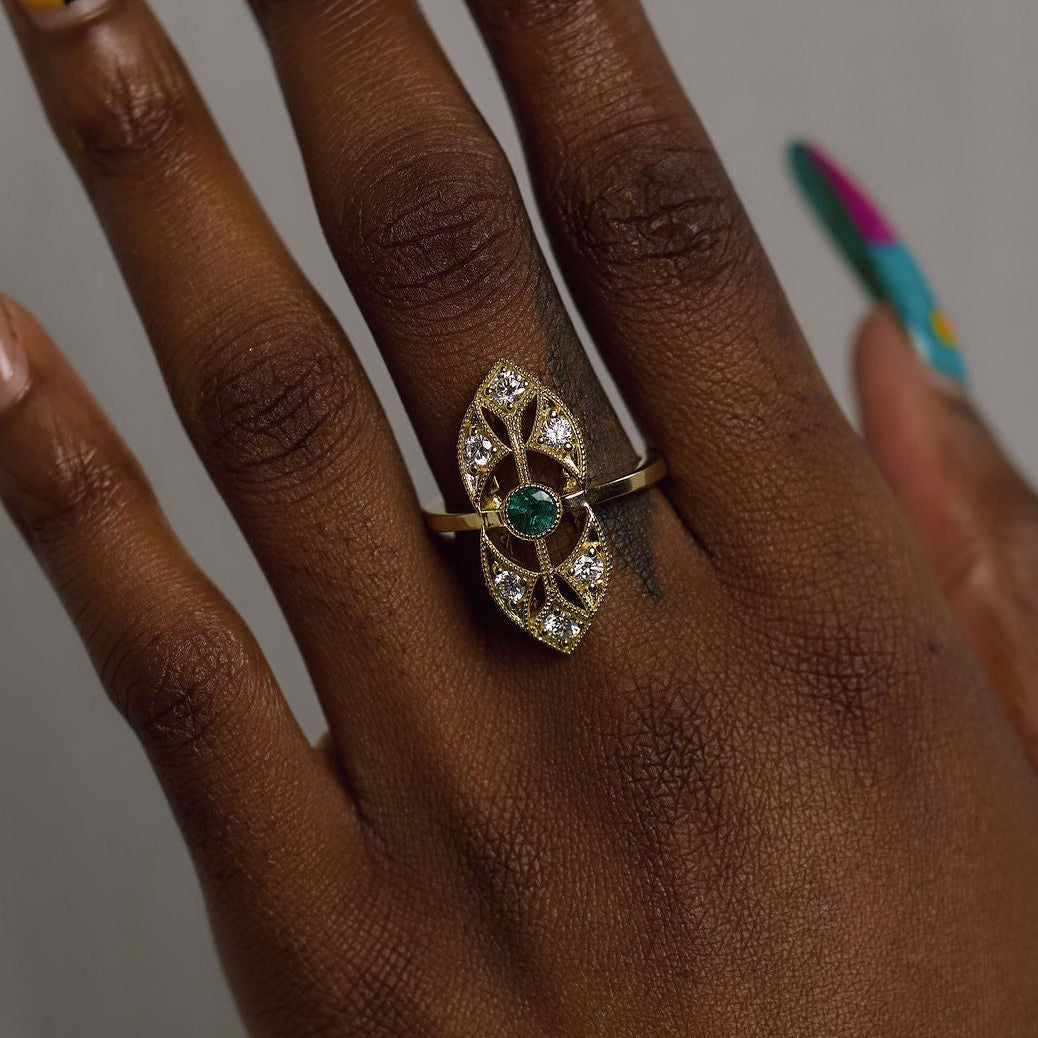 A close-up of a hand with long, colorful manicured nails wearing a gold ring with a green and red central motif and clear stones, set against a plain gray background.