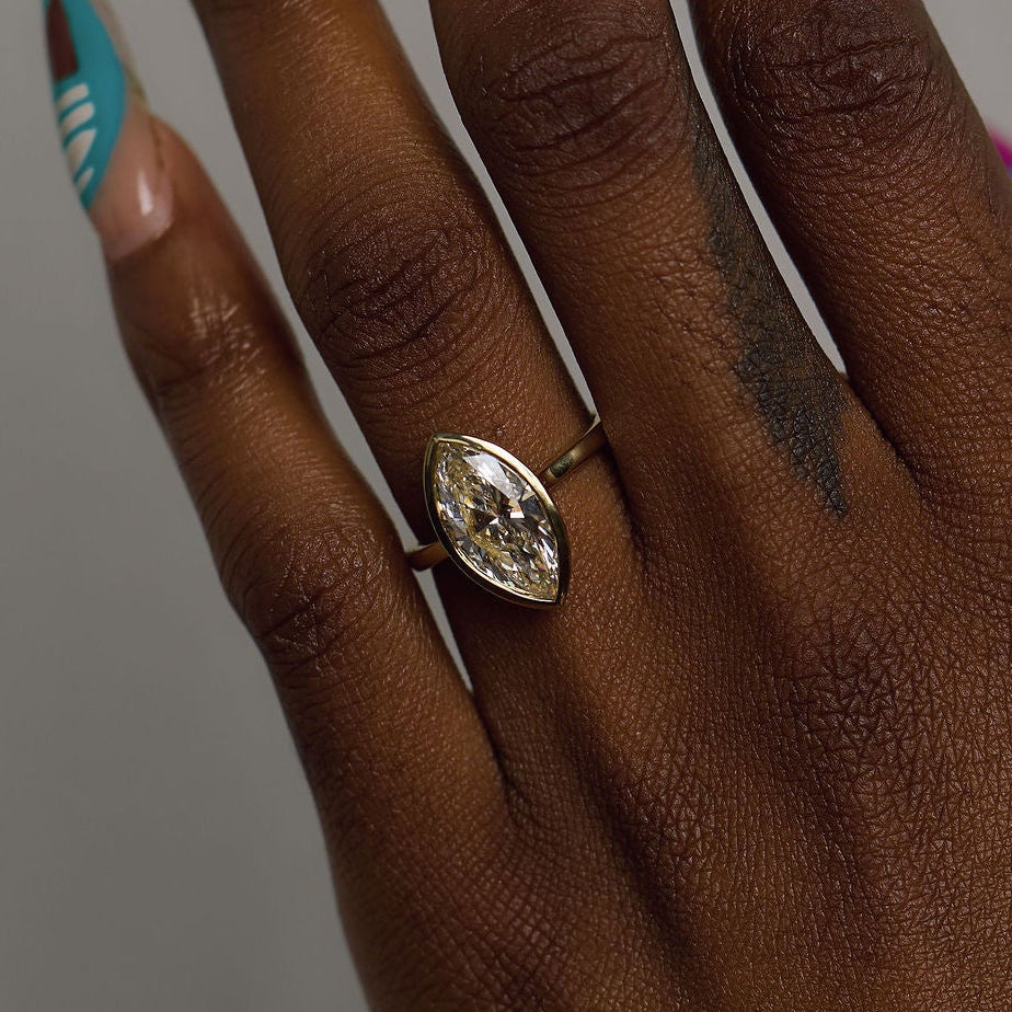 A close-up of a hand wearing a silver-toned ring with a marquise-cut clear gemstone, set against a plain gray background.