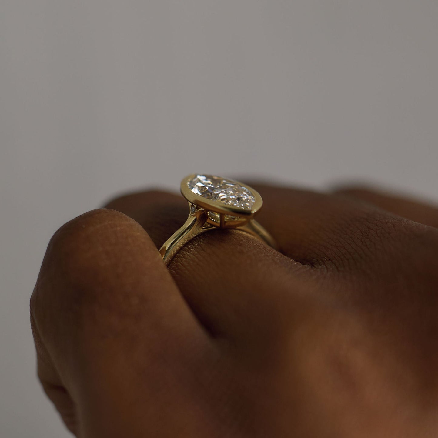 A close-up of a hand wearing a yellow gold ring with a bezel-set round clear gemstone, set against a neutral gray background.