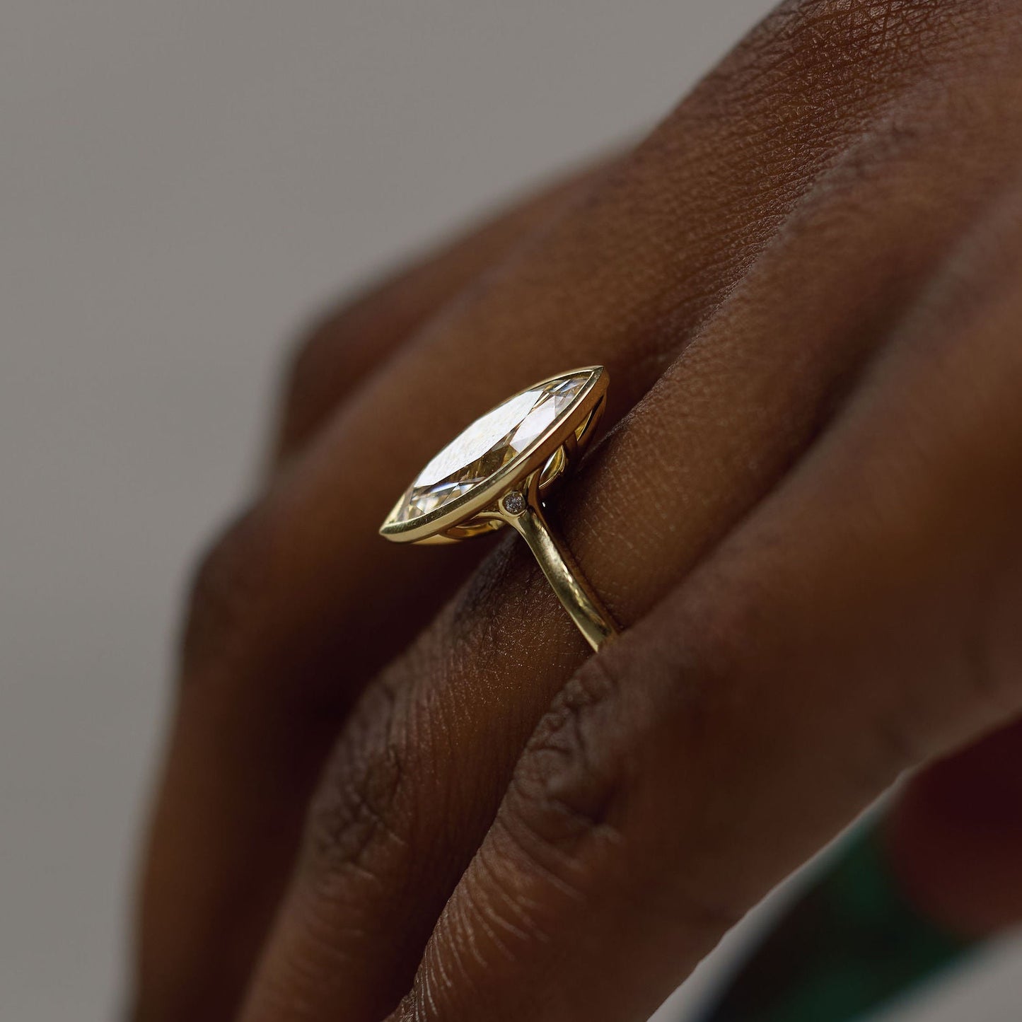 A gold ring with a marquise-cut clear gemstone worn on a person's finger, shown in a close-up shot against a neutral gray background.