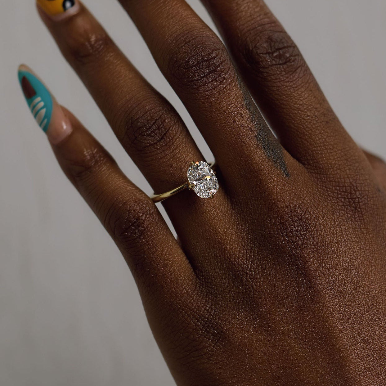 A close-up of a hand with long, colorful manicured nails wearing a delicate gold ring with a large clear oval gemstone, set against a plain light gray background.