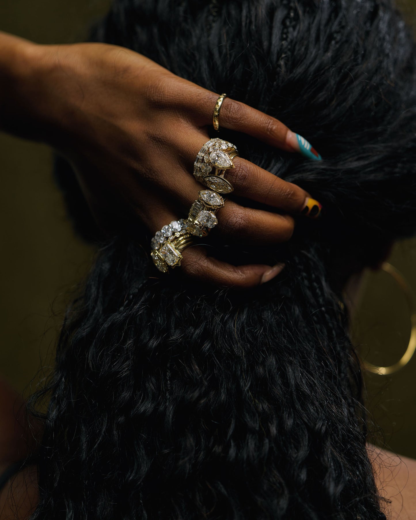 A close-up of a hand with dark skin and long black curly hair, wearing multiple rings, including a large silver-toned ring with clear stones and a smaller gold ring. The hand is holding back the hair against a dark, warm-toned background.
