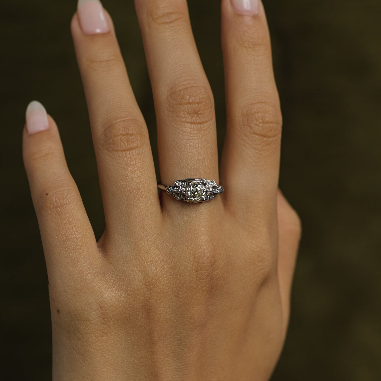 A close-up of a hand with pale pink manicured nails wearing a silver-toned ring with a cluster of small clear stones, set against a dark olive green background.
