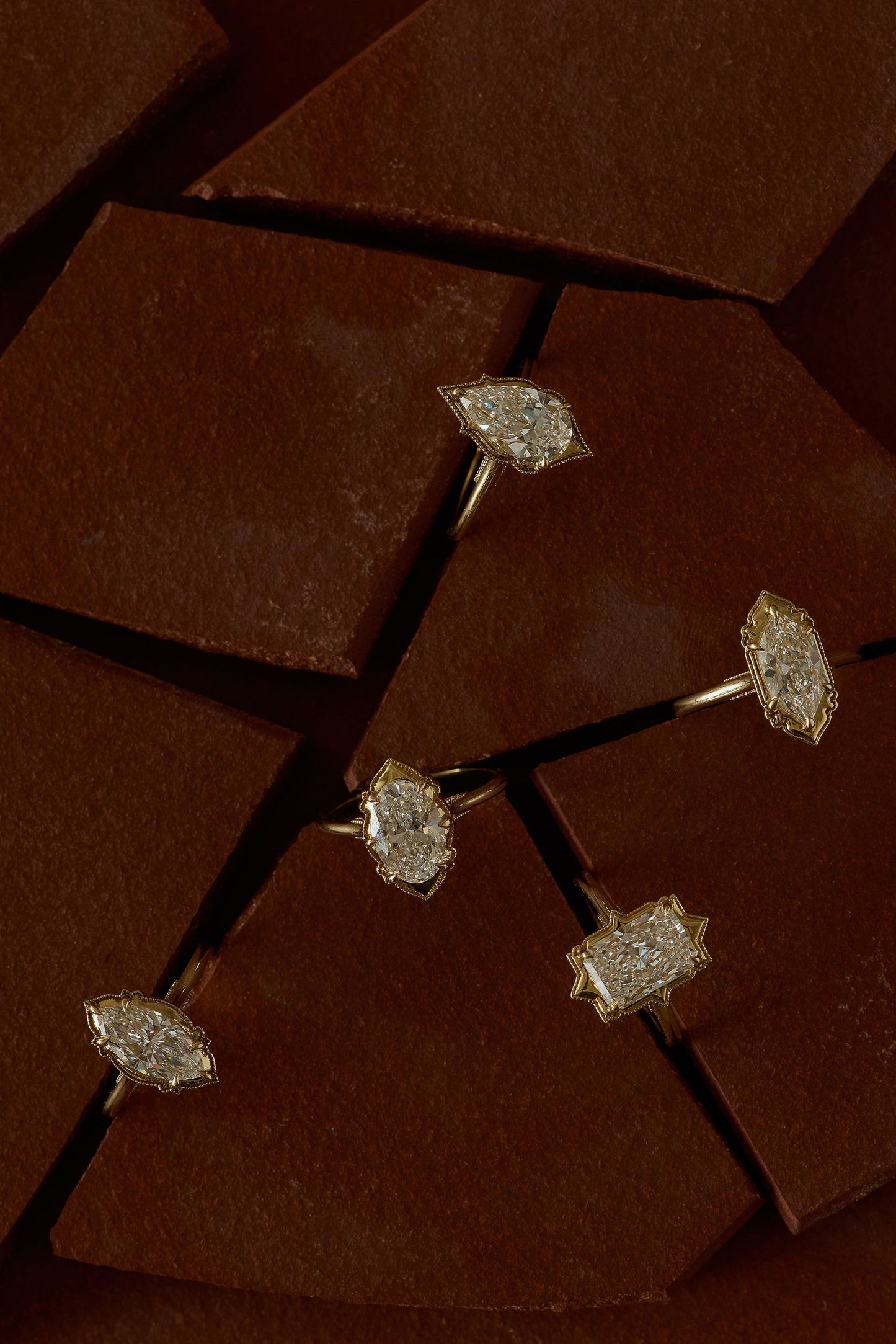 Four gold rings with sparkling white diamonds in different settings, arranged on a dark brown geometric background.