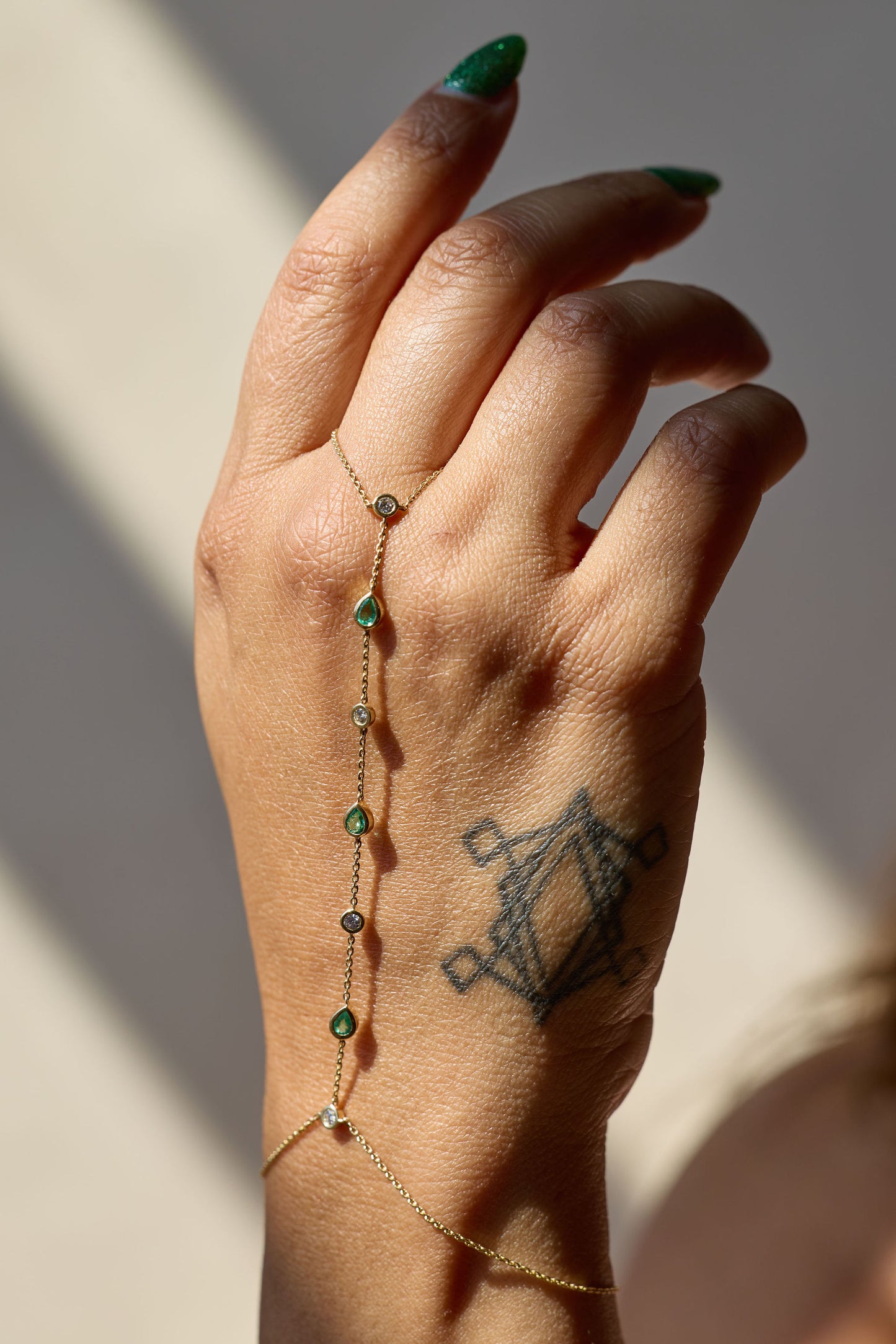 Hand wearing a delicate gold hand chain with emeralds and white diamonds on a neutral background