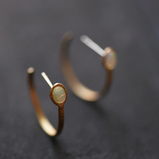 two gold hoop earrings, one in focus, one out of focus, with bezel set opals on a grey background