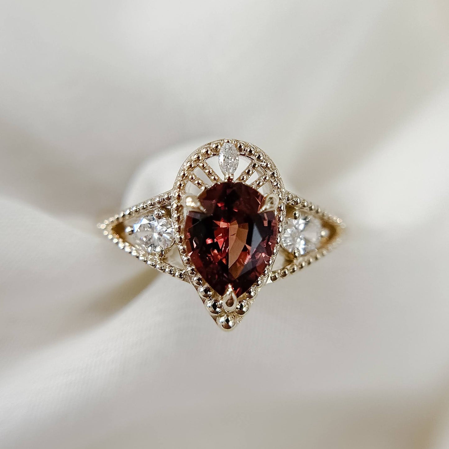 A gold ring featuring a central marquise-cut dark red gemstone surrounded by small clear stones, displayed on a soft white fabric background.