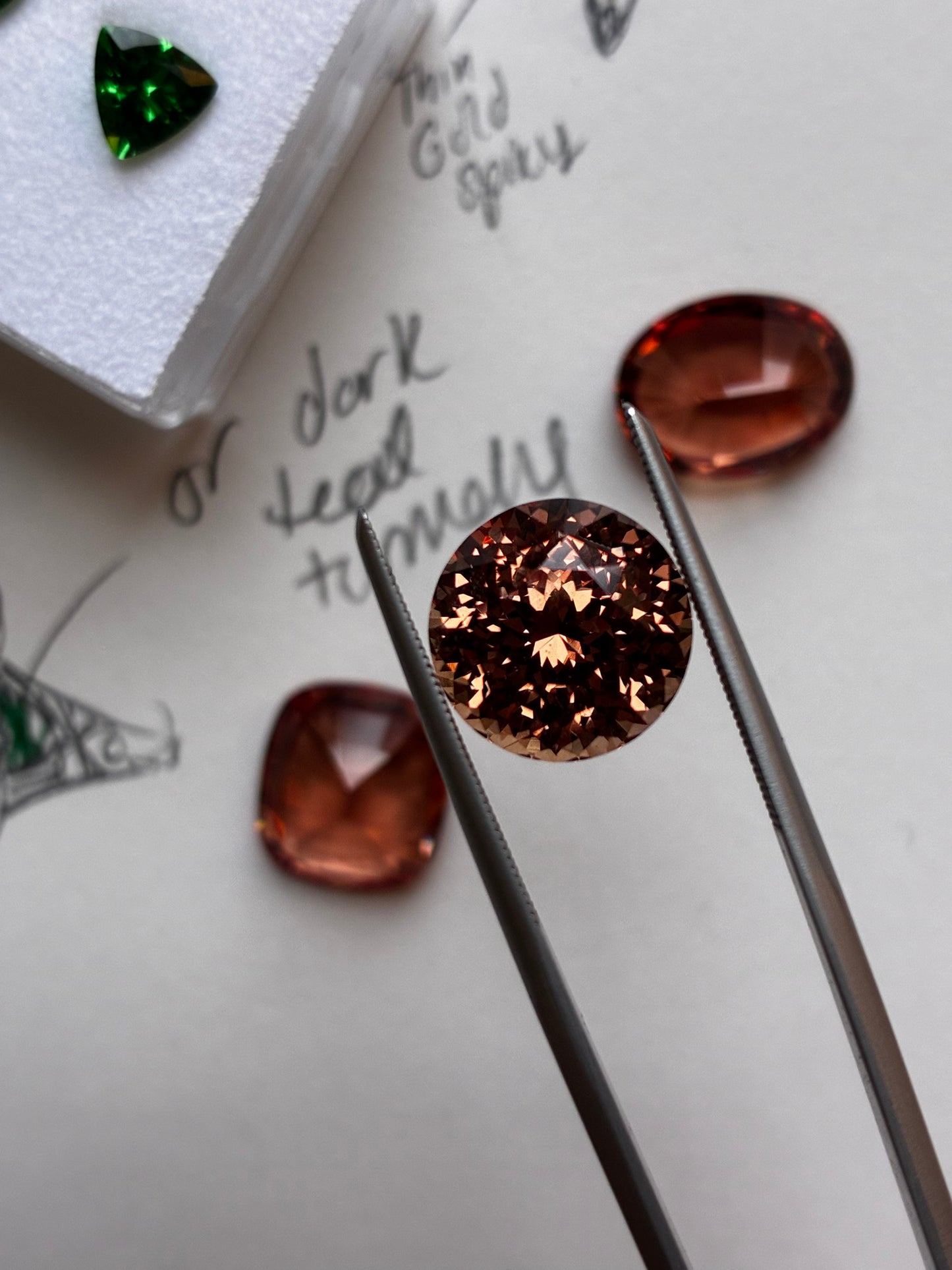 A round brilliant-cut dark copper gemstone held in metal tweezers, with other polished gemstones and handwritten notes on paper in the background.