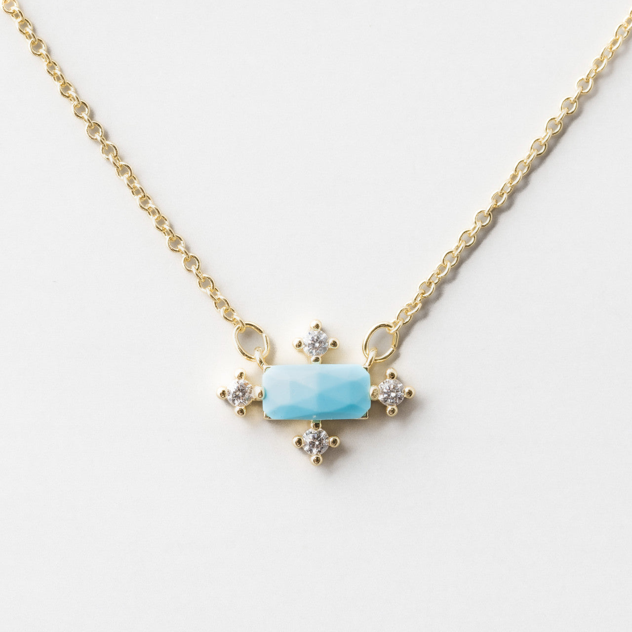 A necklace on a gold chain that has a central turquoise baquette surrounded by Cubic Zirconia on the north, south, east and west points.