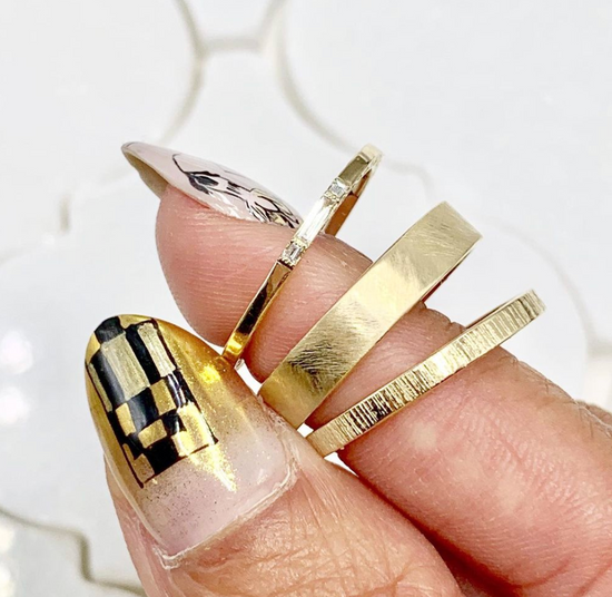 close up of a hand holding three gold bands, one with three small diamonds, on with a brushed texture, and one with birch engraving on a white background
