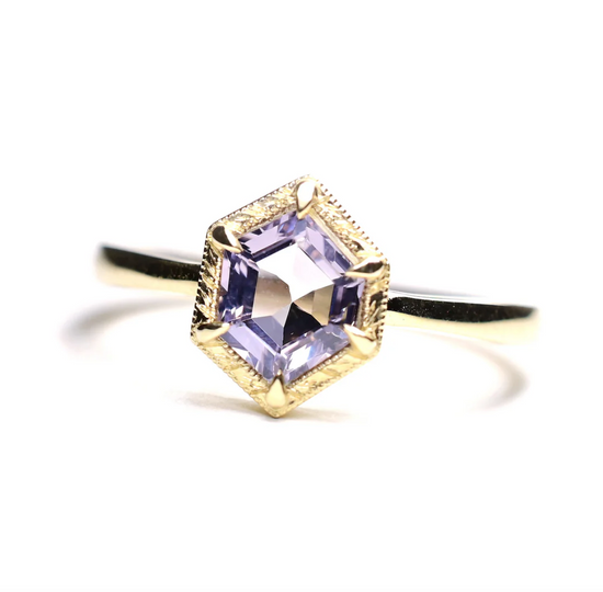 a 14k yellow gold solitaire ring with a lilac purple hexagon shaped spinel with hand engraved gold details around the center stone, sitting on a white background