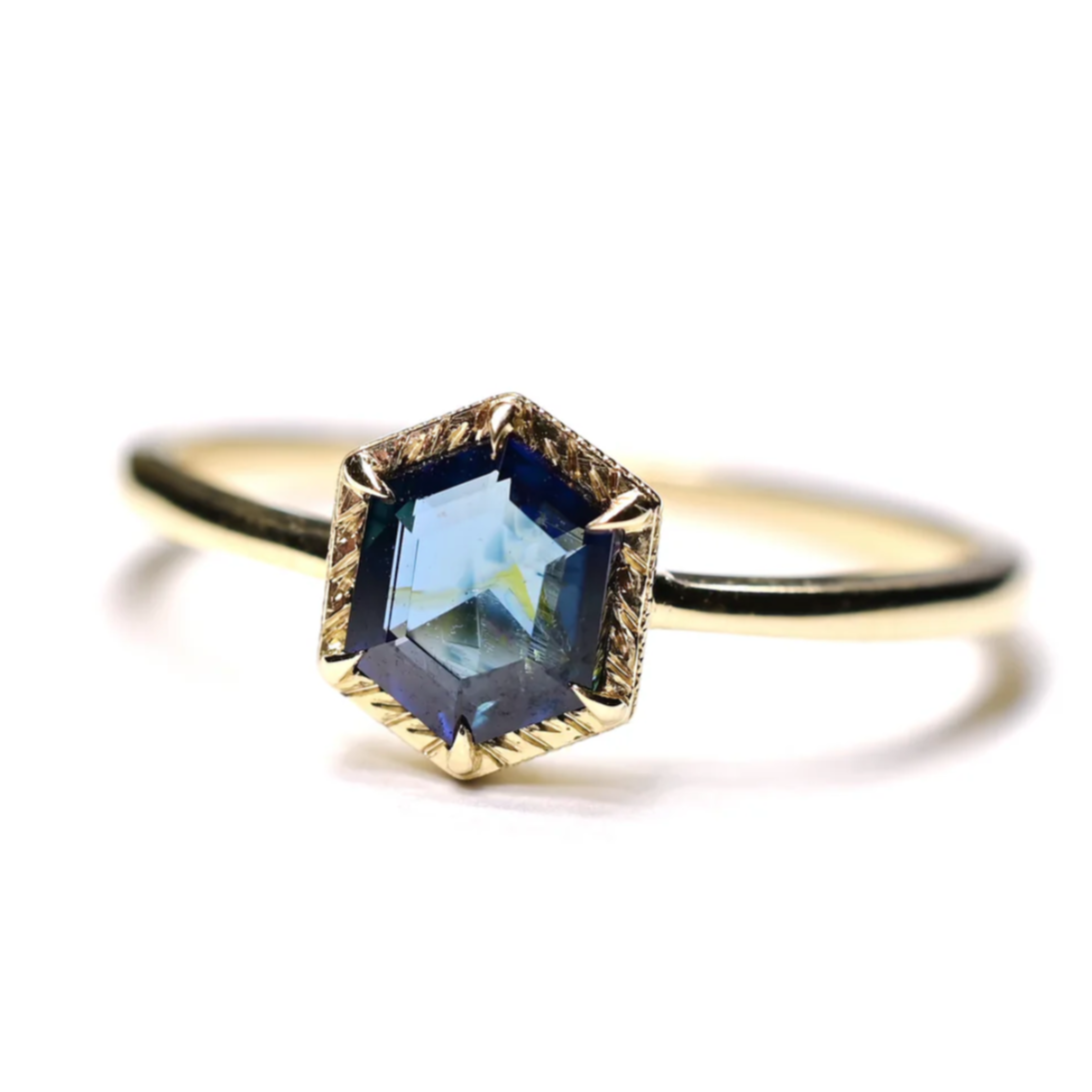 a 14k yellow gold solitaire ring with a hexagon blue spinel center stone with engraved gold details around the stone, sitting on a white background