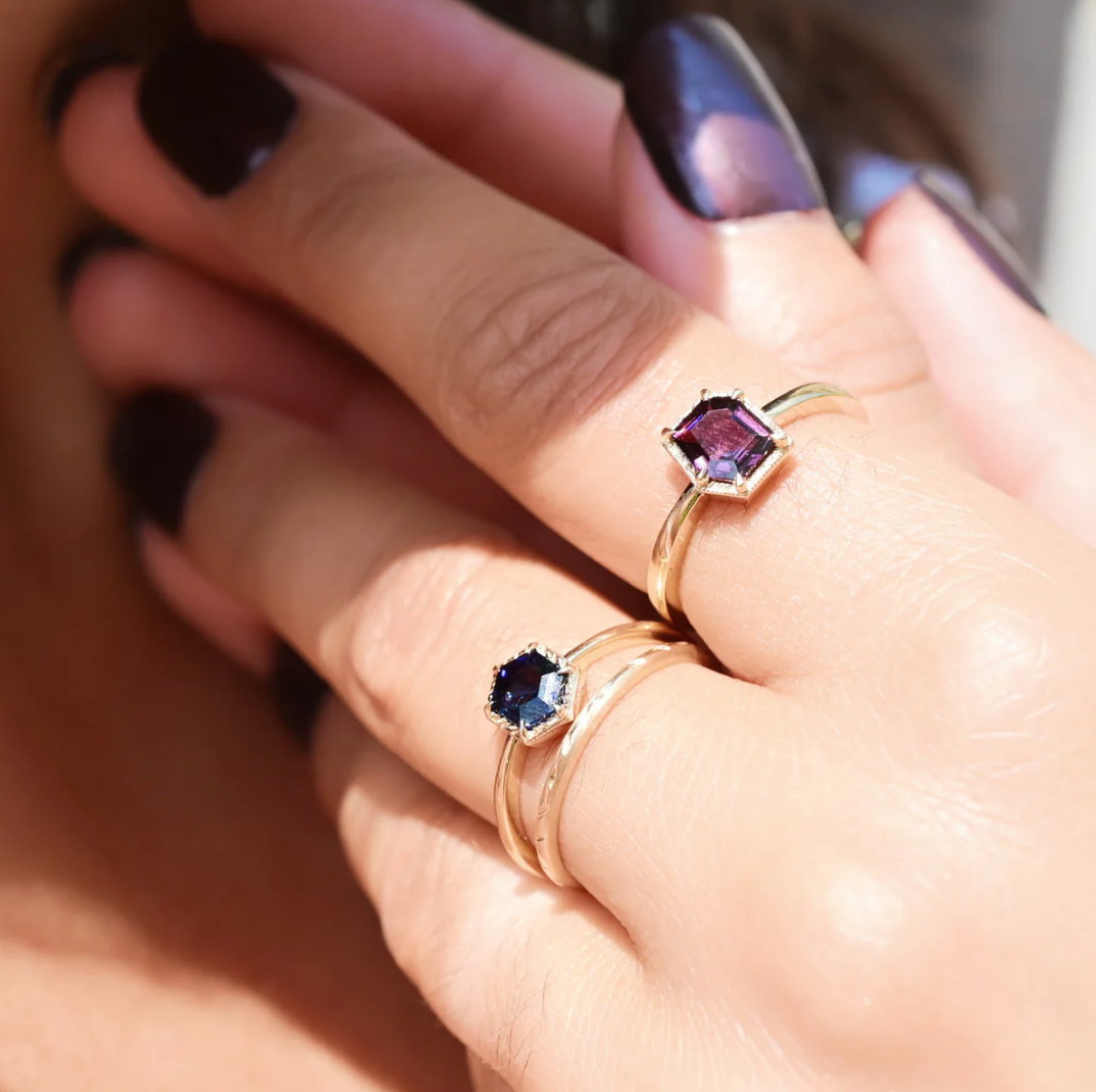 close up of a model wearing the hexagon blue spinel solitaire ring on her middle finger stacked with a straight gold band and a purple spinel solitaire ring on her pointer finger