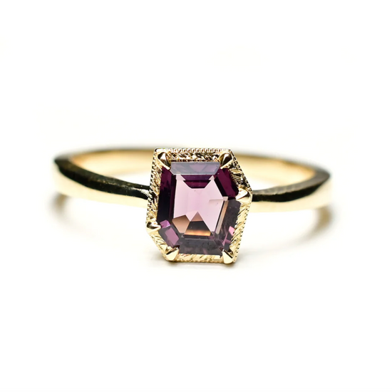 a 14k yellow gold solitaire ring with a fancy cut purple spinel center stone with engraved gold details around the stone, sitting on a white background