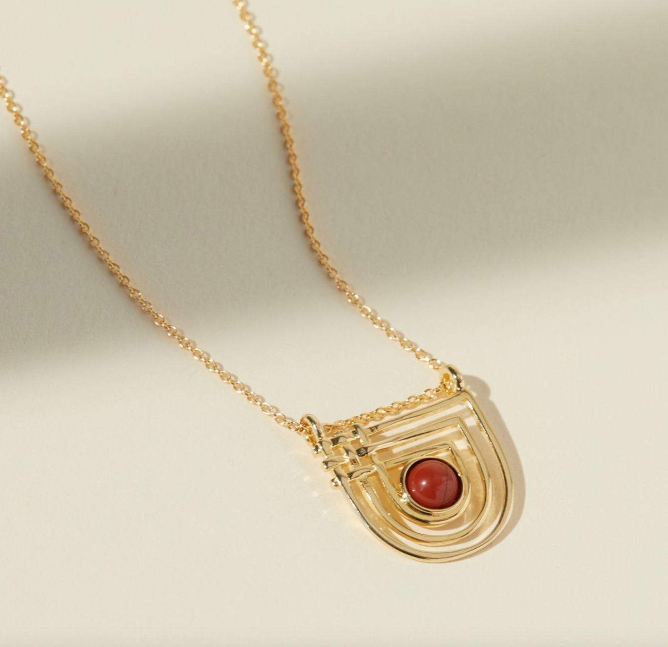 curved, crosshatched pendant necklace with a red jasper center stone on a beige background
