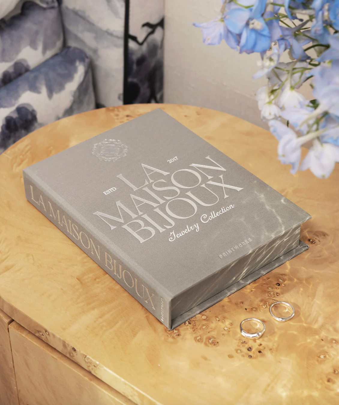 Gray jewelry box titled 'La Maison Bijoux' on a wooden surface with flowers in the background