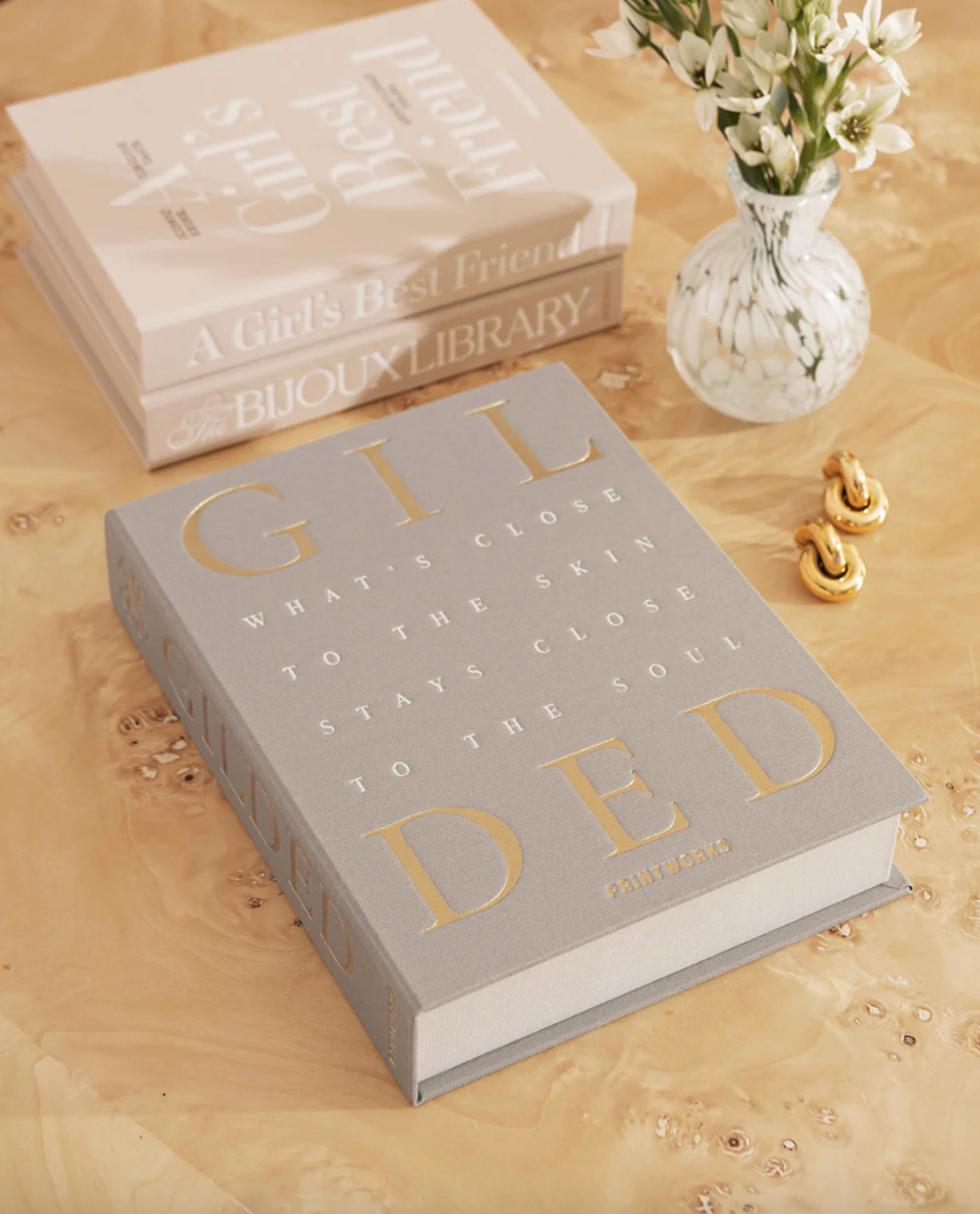 Gray jewelry box titled 'GILDED' on a wooden surface with a vase of flowers in the background