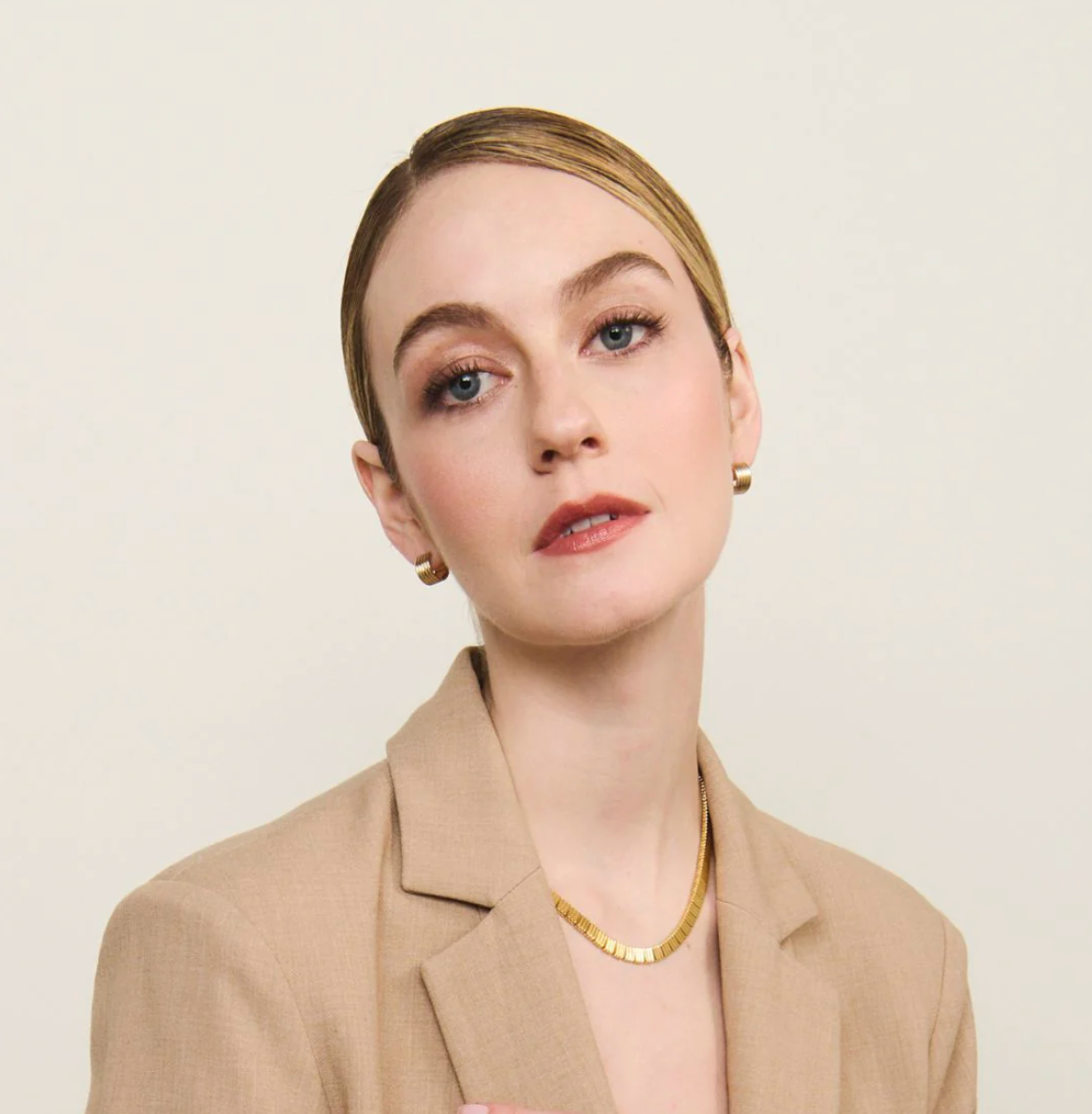 model wearing gold hoop earrings, a gold chain necklace and beige blazer on grey background