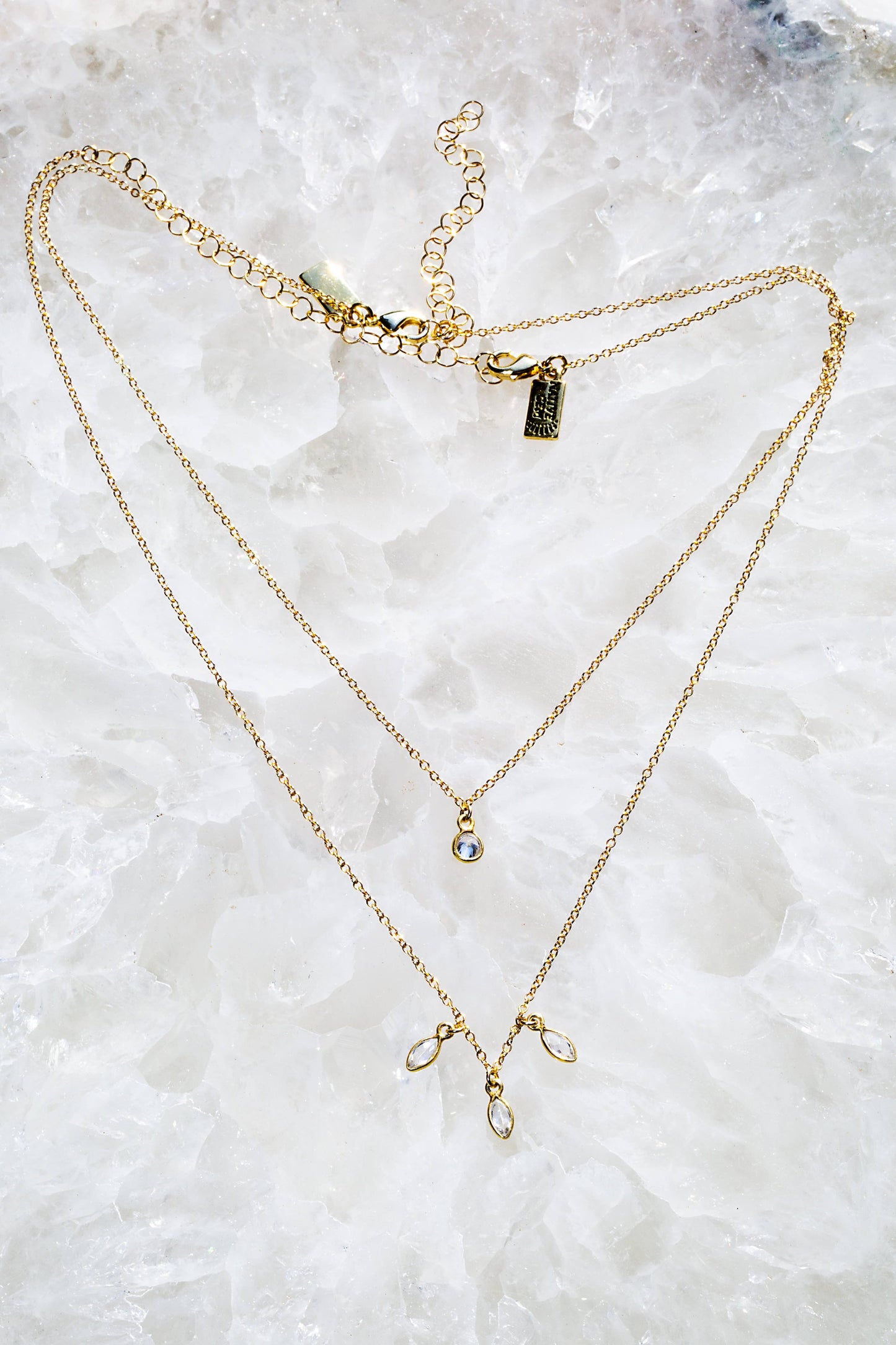 Two gold necklaces with small pendants on a white stone background
