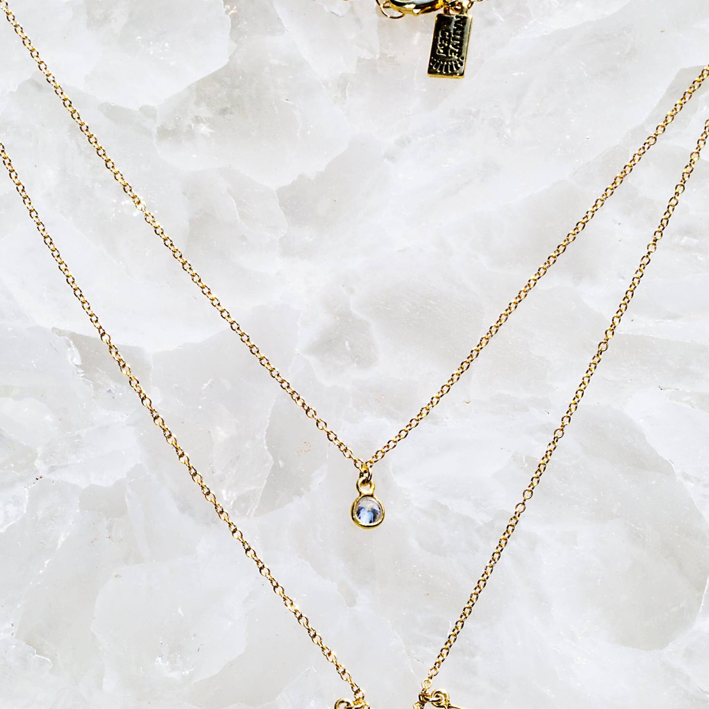Two gold necklaces with small pendants on a white stone background