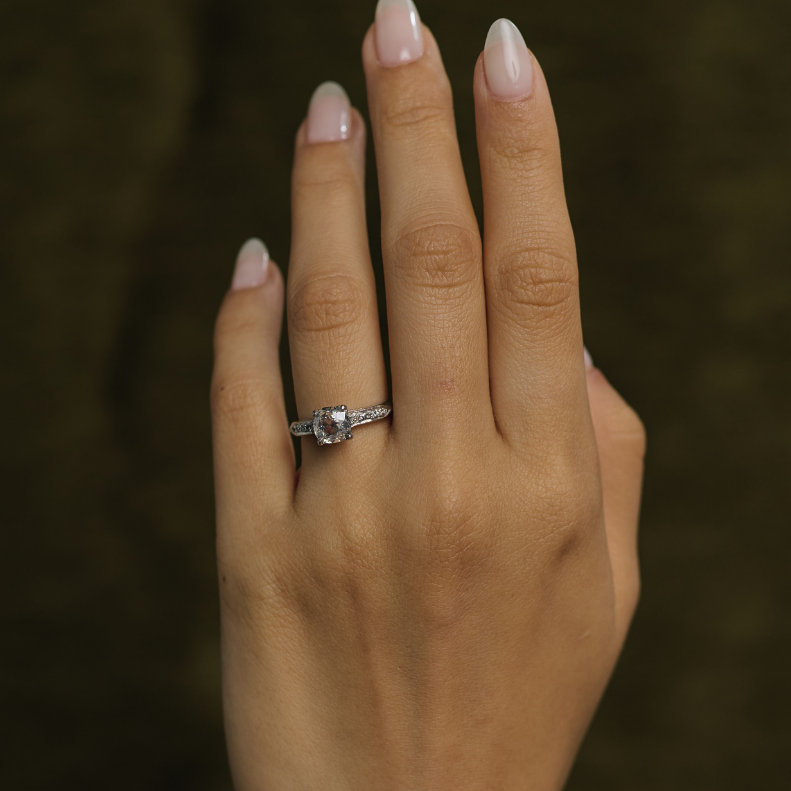 A close-up of a hand wearing a silver ring with a clear center stone, set against a dark olive green background.