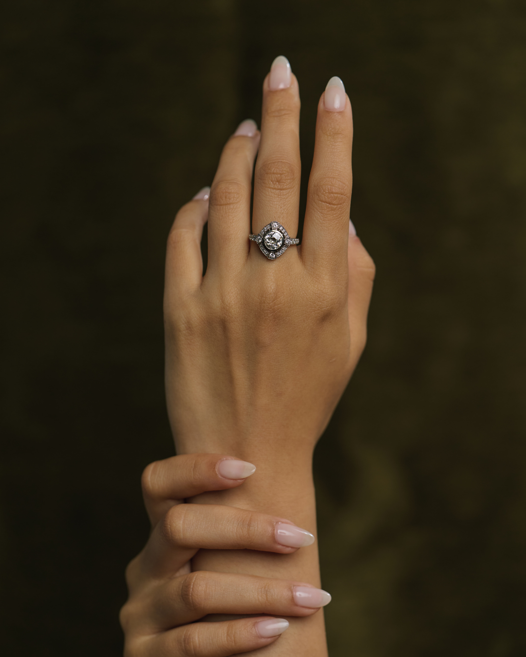 A close-up of a hand wearing an antique-style silver ring with a central clear gemstone, set against a dark, textured olive-green background.