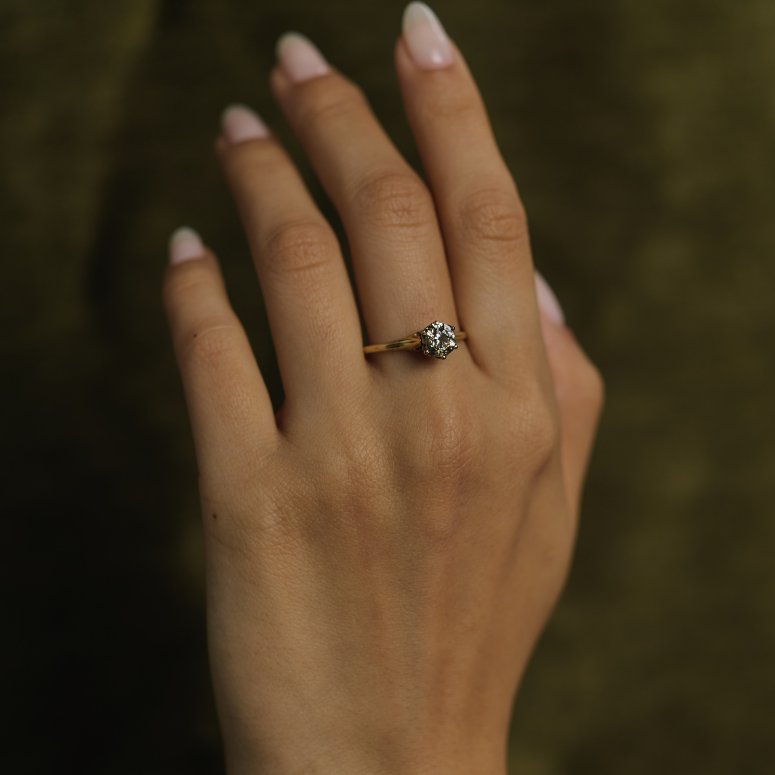 A close-up of a hand wearing a delicate gold ring with a small round clear gemstone, set against a dark olive green textured background.