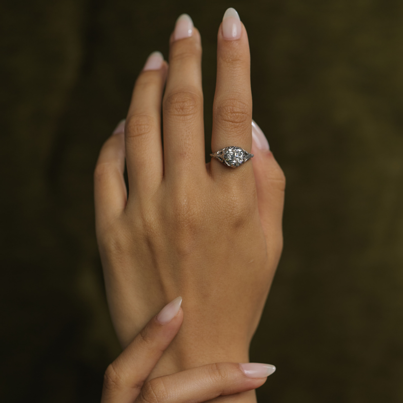 A close-up of a person's hands with manicured nails, wearing a silver-toned ring with a clear heart-shaped gemstone on one finger, set against a dark olive green textured background.