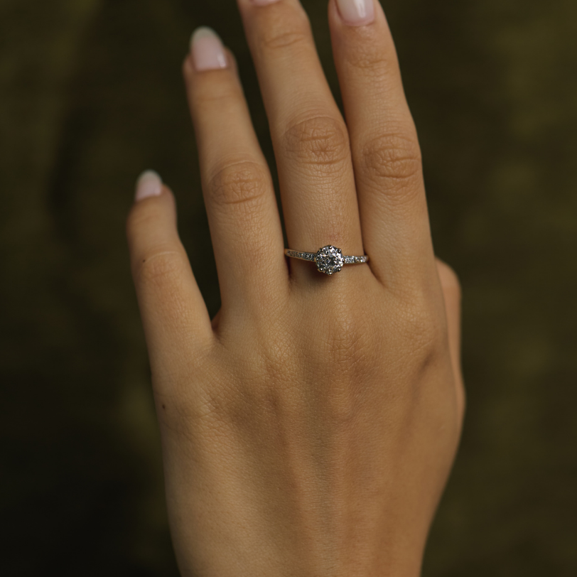 A close-up of a hand with light pink manicured nails wearing a silver-toned ring with a small clear central gemstone, set against a dark olive green textured background.