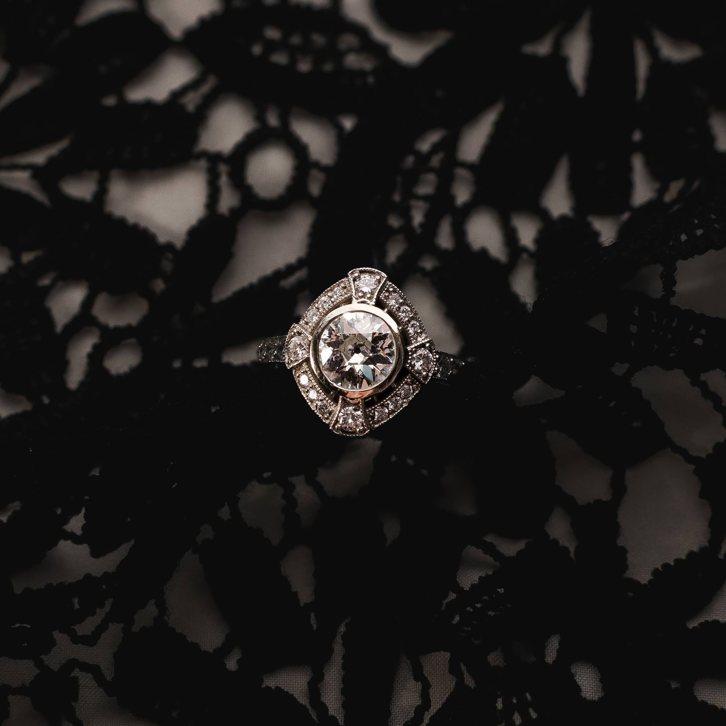 A silver-toned diamond ring with a square-shaped center stone and surrounding pavé setting, resting on a black lace fabric background.