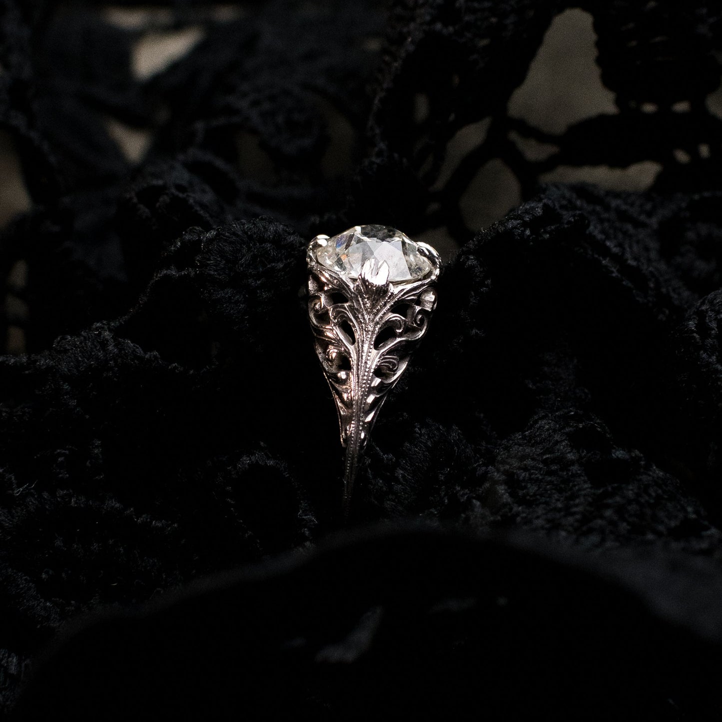 An ornate silver-toned ring with a clear round gemstone, displayed against a dark black lace fabric background.