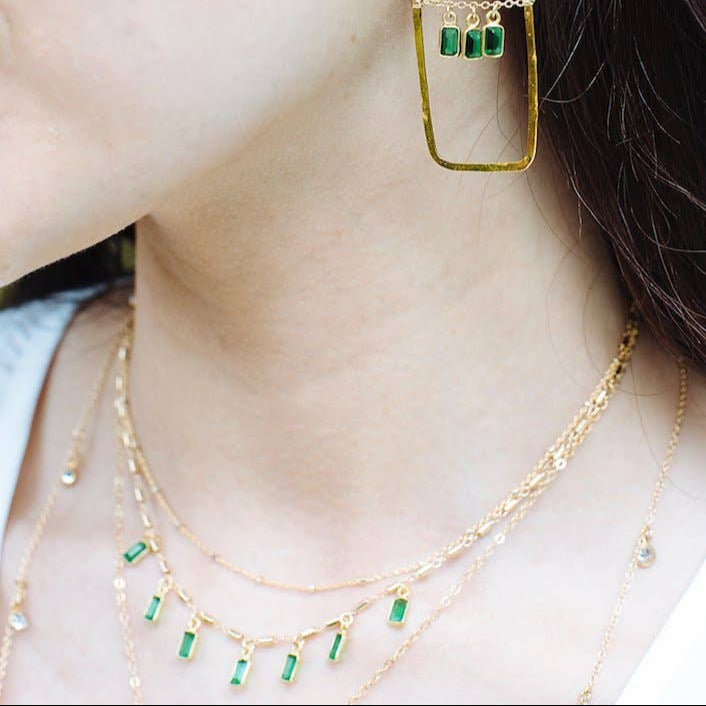 Close-up of a person wearing gold hoop earrings with green gemstones and matching necklaces.