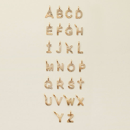 textured gold initial charms displayed in alphabetical order a through z on a beige background
