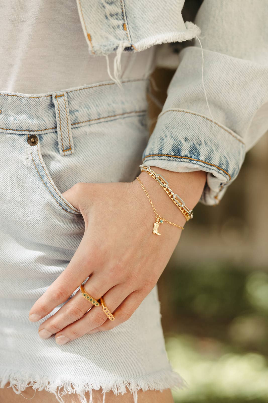 Hand wearing gold bracelets and a ring with a blurred natural background