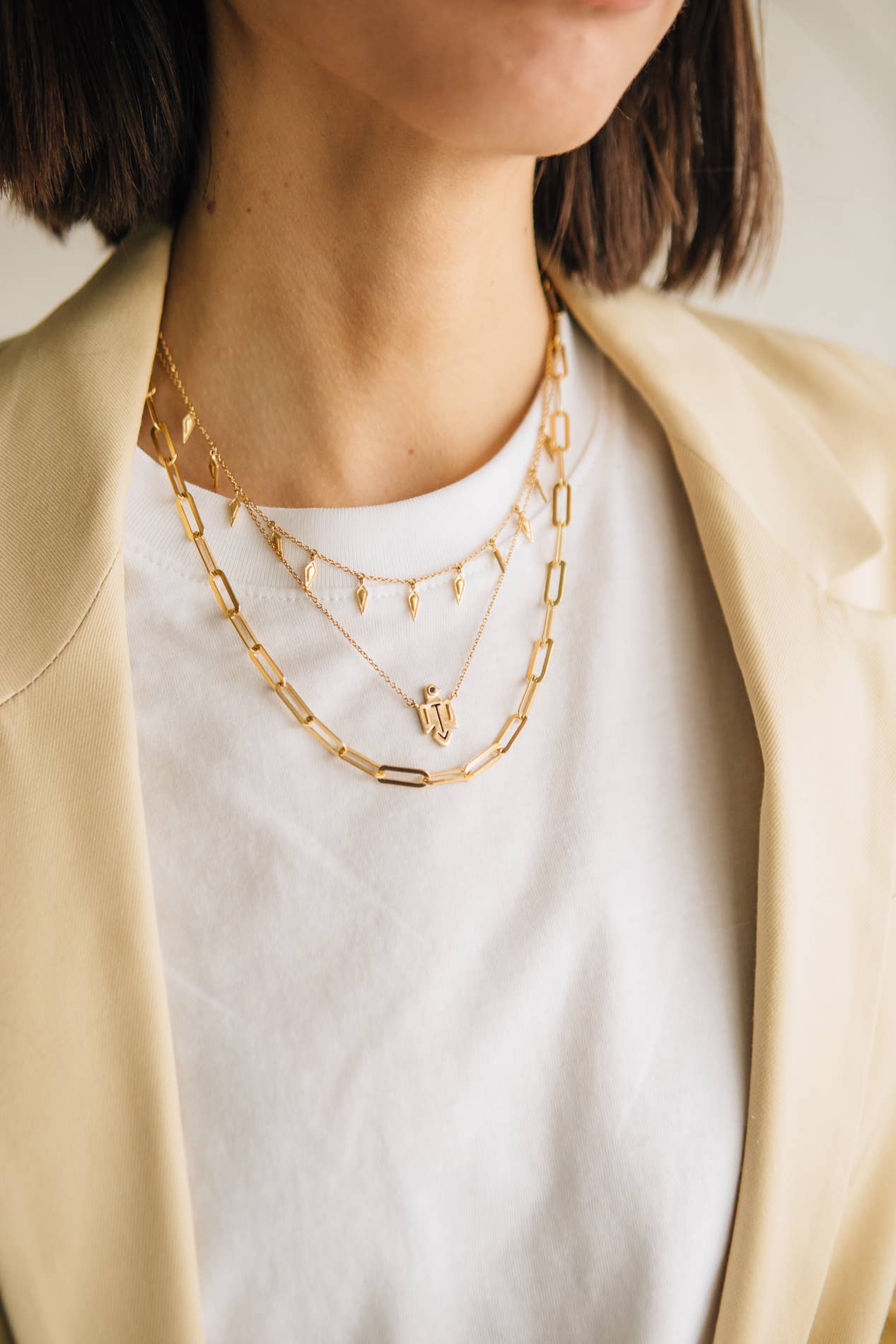 Person wearing a white shirt and gold chain necklaces with a beige jacket.
